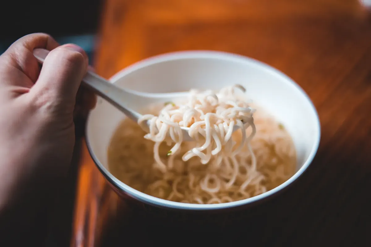 A hand holding a spoonful of instant noodles over a bowl, perfect for food-related designs.